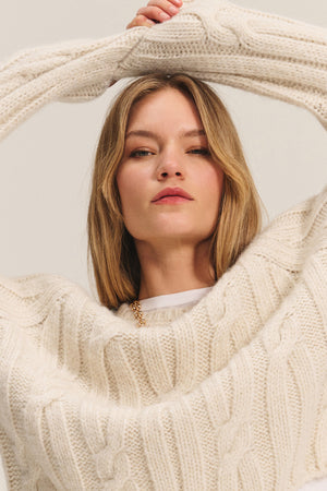 A woman poses with her arms raised above her head, wearing the MARINA SWEATER by Velvet by Graham & Spencer—a cream-colored cable knit with subtle metallic threading—against a plain background.
