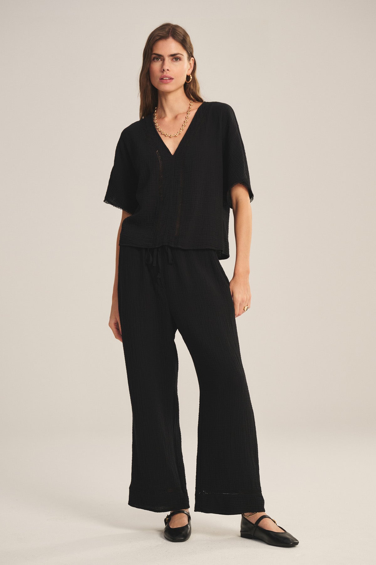 A woman faces forward against a plain light background, wearing the CALIA PANT by Velvet by Graham & Spencer—a loose black cotton gauze pant with a drawstring waist—paired with a matching V-neck top, black flats, and a gold necklace.