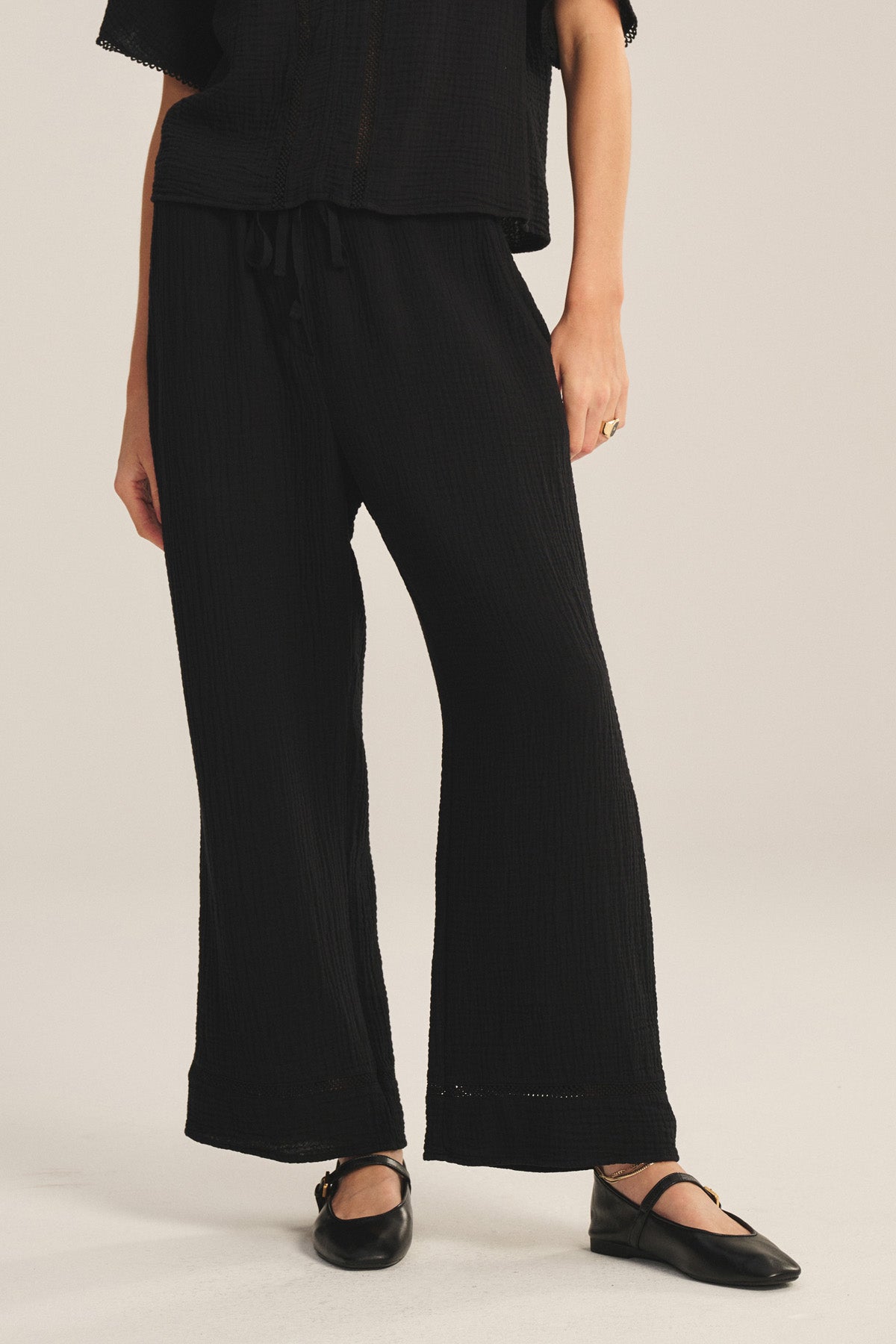 A person wears the CALIA PANT by Velvet by Graham & Spencer—loose-fitting black cotton gauze pants with a drawstring waist—paired with a matching top and black flats, standing on a neutral background.-41296660168897