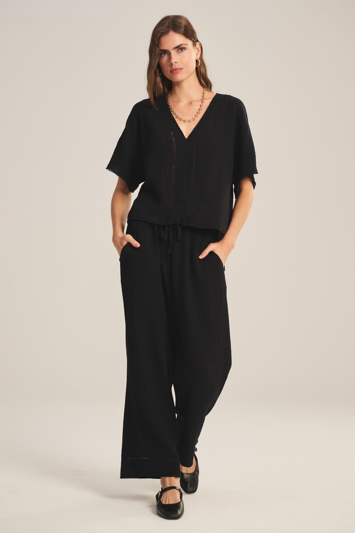 A woman stands against a plain light background wearing Velvet by Graham & Spencer's loose-fitting black CALIA PANT in cotton gauze with a drawstring waist and pockets, paired with a matching V-neck top.