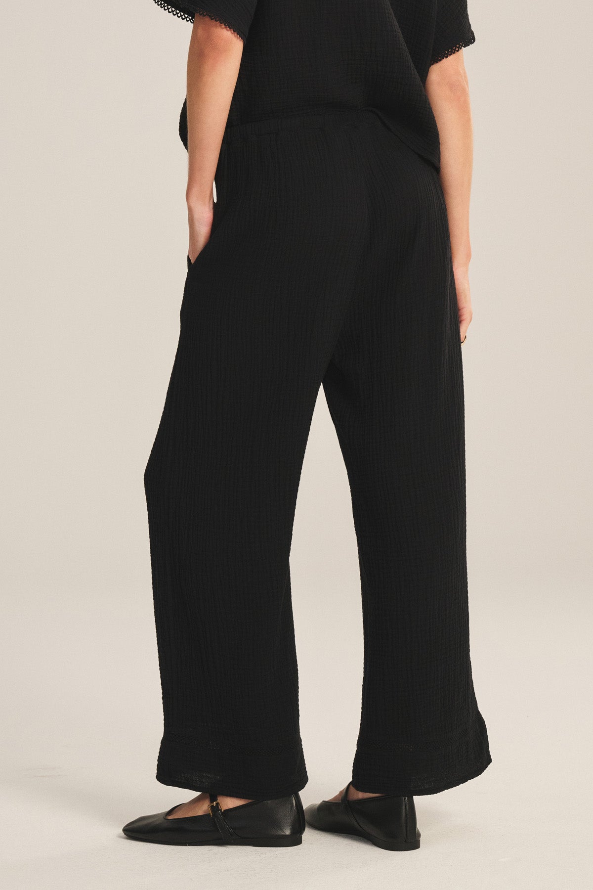A person wearing the CALIA PANT by Velvet by Graham & Spencer—black cotton gauze wide-leg pants with a drawstring waist—and black flat shoes, stands with hands in pockets, facing away from the camera.