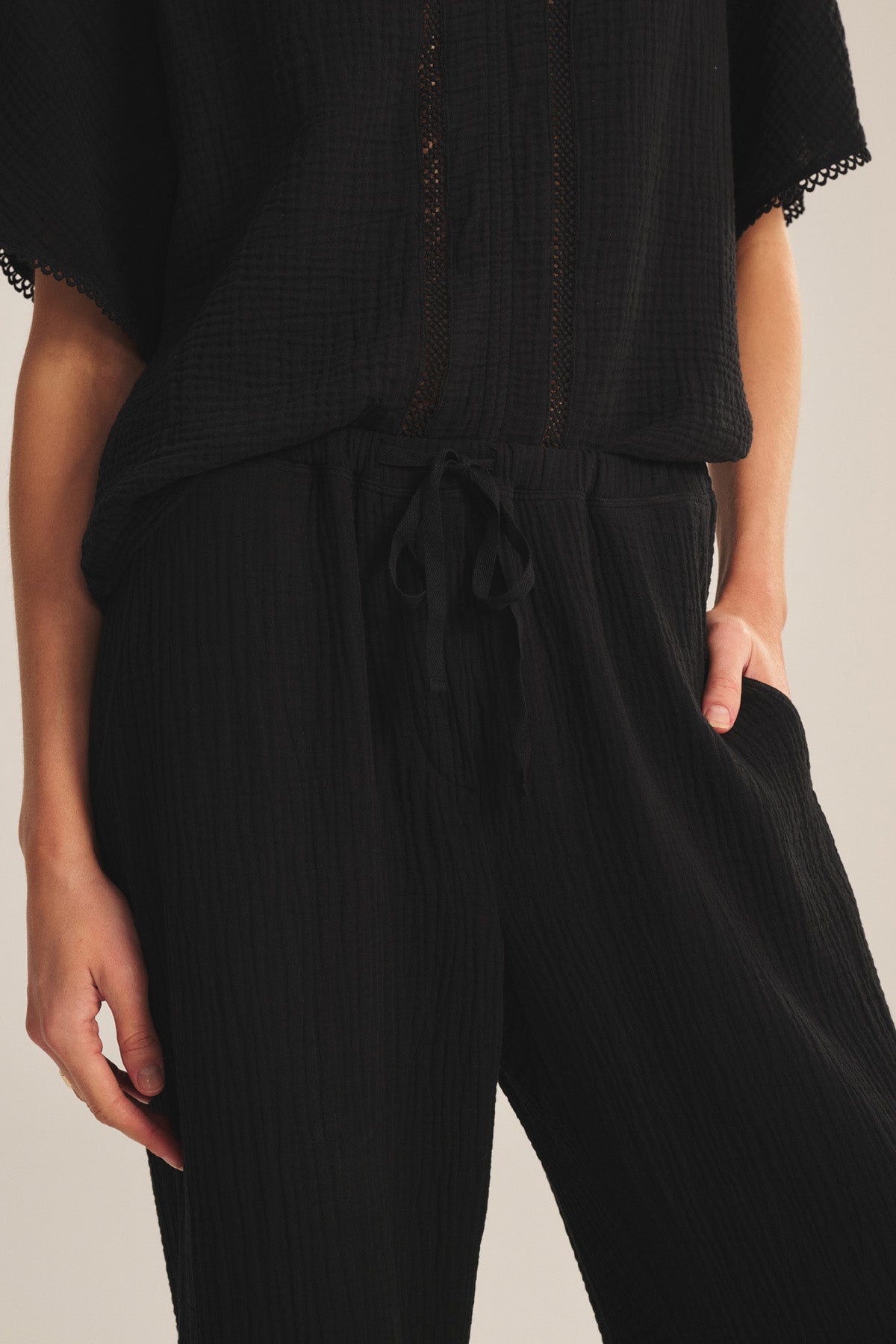 A person, hand in pocket, is photographed from shoulder to thigh wearing the CALIA PANT by Velvet by Graham & Spencer—a black textured cotton gauze style with a drawstring waist.