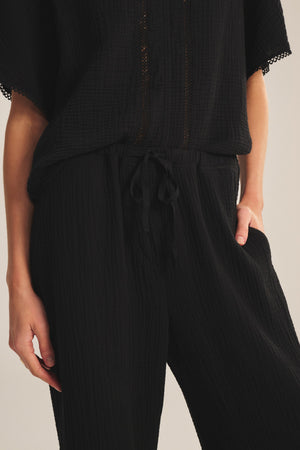 A person, hand in pocket, is photographed from shoulder to thigh wearing the CALIA PANT by Velvet by Graham & Spencer—a black textured cotton gauze style with a drawstring waist.