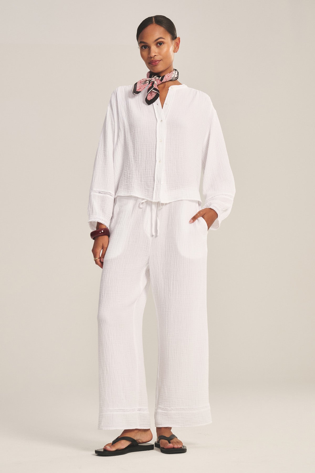A woman stands in a studio wearing a loose white button-up top, Velvet by Graham & Spencer’s CALIA PANT in cotton gauze with drawstring elastic waist, black sandals, and a pink patterned scarf around her neck.-41128033910977