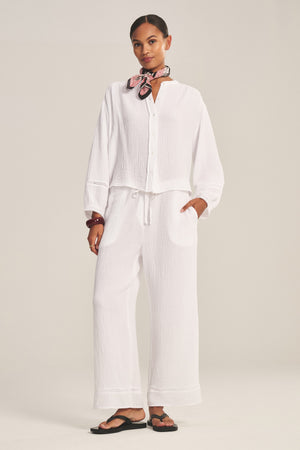 A woman stands in a studio wearing a loose white button-up top, Velvet by Graham & Spencer’s CALIA PANT in cotton gauze with drawstring elastic waist, black sandals, and a pink patterned scarf around her neck.