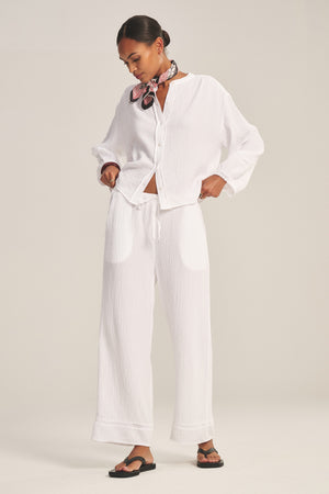 A woman stands in a relaxed pose wearing a white button-up shirt, Velvet by Graham & Spencer’s CALIA PANT in cotton gauze with a drawstring elastic waist, black sandals, and a patterned neck scarf.
