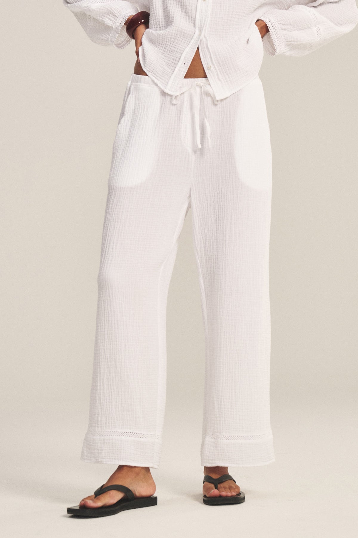 A person wears the CALIA PANT by Velvet by Graham & Spencer—loose white cotton gauze pants with a drawstring waist—styled with a matching button-up top and black sandals, standing against a plain background.