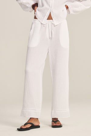A person wears the CALIA PANT by Velvet by Graham & Spencer—loose white cotton gauze pants with a drawstring waist—styled with a matching button-up top and black sandals, standing against a plain background.