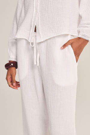 Model wears the CALIA PANT by Velvet by Graham & Spencer—a cotton gauze drawstring pant—paired with a white textured long-sleeve top featuring lace cut-out trim on the sleeves, one hand casually in their pocket.