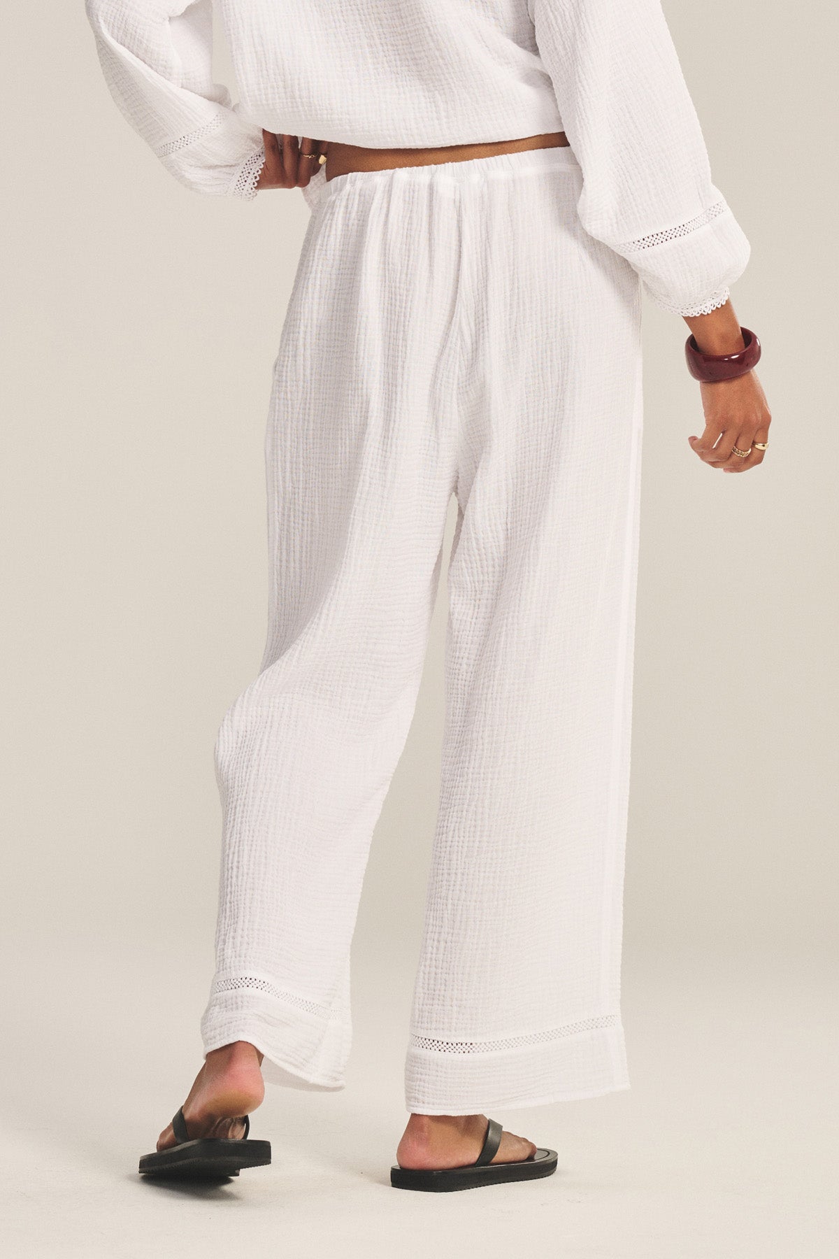 Model wears the CALIA PANT by Velvet by Graham & Spencer—loose white cotton gauze pants with a drawstring elastic waist—paired with a matching lace cut-out top and sandals, shown from the back against a light background.