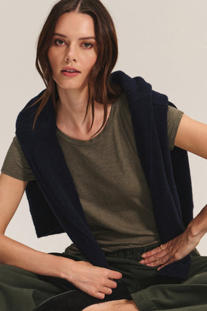 A woman with long brown hair wears the ODELIA TEE by Velvet by Graham & Spencer in olive green, featuring a classic crew neckline, styled with dark green pants and a navy sweater over her shoulders—a true wardrobe essential.