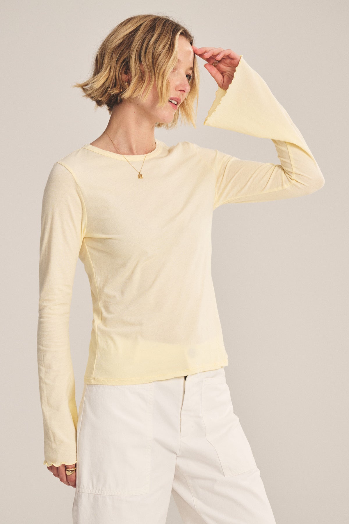 A woman in the light yellow CAMILLE TEE by Velvet by Graham & Spencer with bell sleeves and white pants stands facing left, raising her right hand to her forehead.-41008168075457