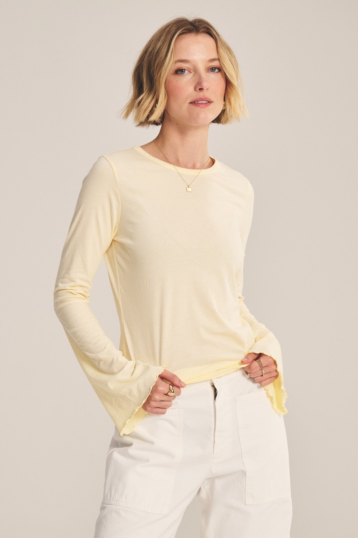 A woman with short blonde hair wears the CAMILLE TEE by Velvet by Graham & Spencer—a light yellow fitted top with bell sleeves—paired with white pants, standing hands-in-pockets against a plain backdrop.-41008168042689