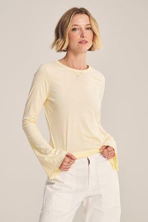 A woman with short blonde hair wears the CAMILLE TEE by Velvet by Graham & Spencer—a light yellow fitted top with bell sleeves—paired with white pants, standing hands-in-pockets against a plain backdrop.