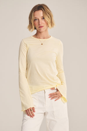 A person with short blonde hair wears the CAMILLE TEE by Velvet by Graham & Spencer, a light yellow fitted top with bell sleeves, paired with white pants, standing against a plain light background.