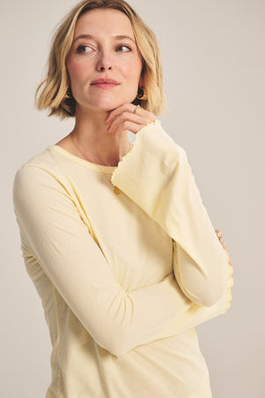 A woman with short blonde hair wears the CAMILLE TEE by Velvet by Graham & Spencer in light yellow, looking slightly to the side with her hand resting near her chin.