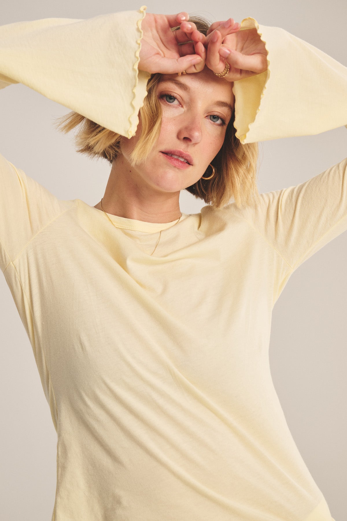 A person with short blonde hair wears the CAMILLE TEE by Velvet by Graham & Spencer—a light yellow fitted top with bell sleeves—and holds their hands above their forehead, looking at the camera.