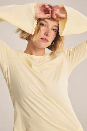 A person with short blonde hair wears the CAMILLE TEE by Velvet by Graham & Spencer—a light yellow fitted top with bell sleeves—and holds their hands above their forehead, looking at the camera.