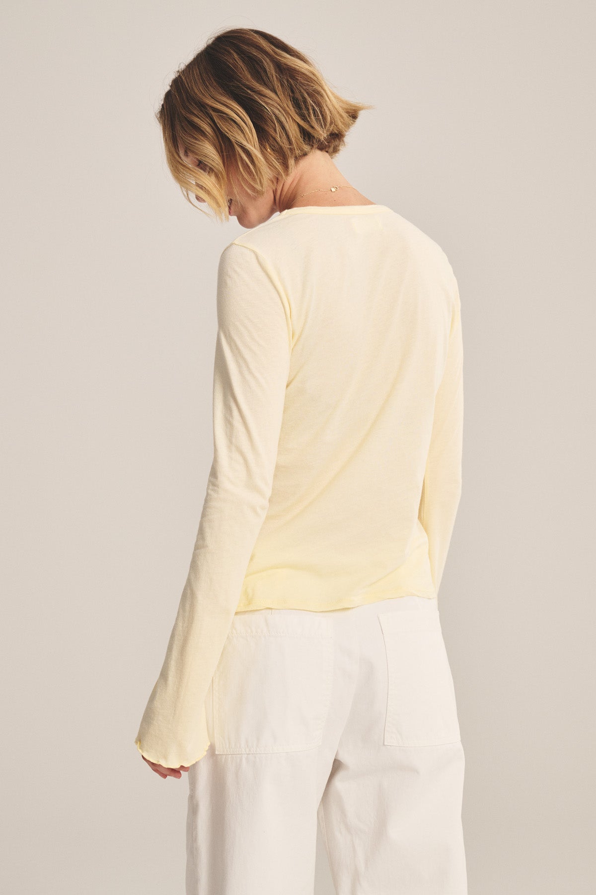 A person with short hair stands with their back to the camera, wearing the CAMILLE TEE by Velvet by Graham & Spencer in pale yellow and white pants.-41008167780545