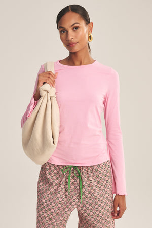 A woman stands against a neutral background wearing the CAMILLE TEE by Velvet by Graham & Spencer in light pink, paired with patterned pants, gold earrings, and a beige knotted cloth bag over her shoulder.