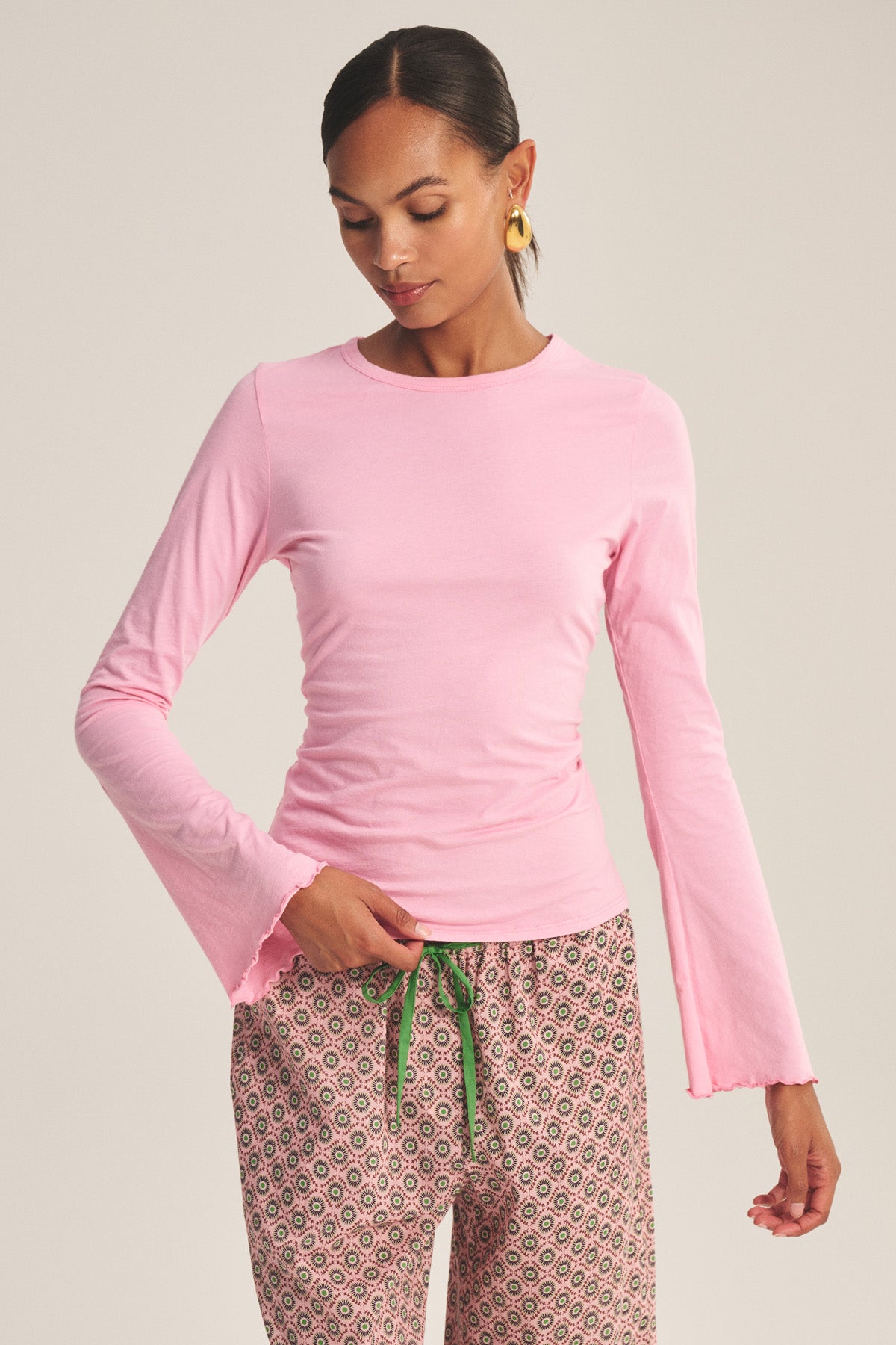 A woman wears the CAMILLE TEE by Velvet by Graham & Spencer, a long-sleeve pink top with scalloped cuffs, paired with patterned pants featuring a green drawstring, standing against a plain light background.-41296690577601