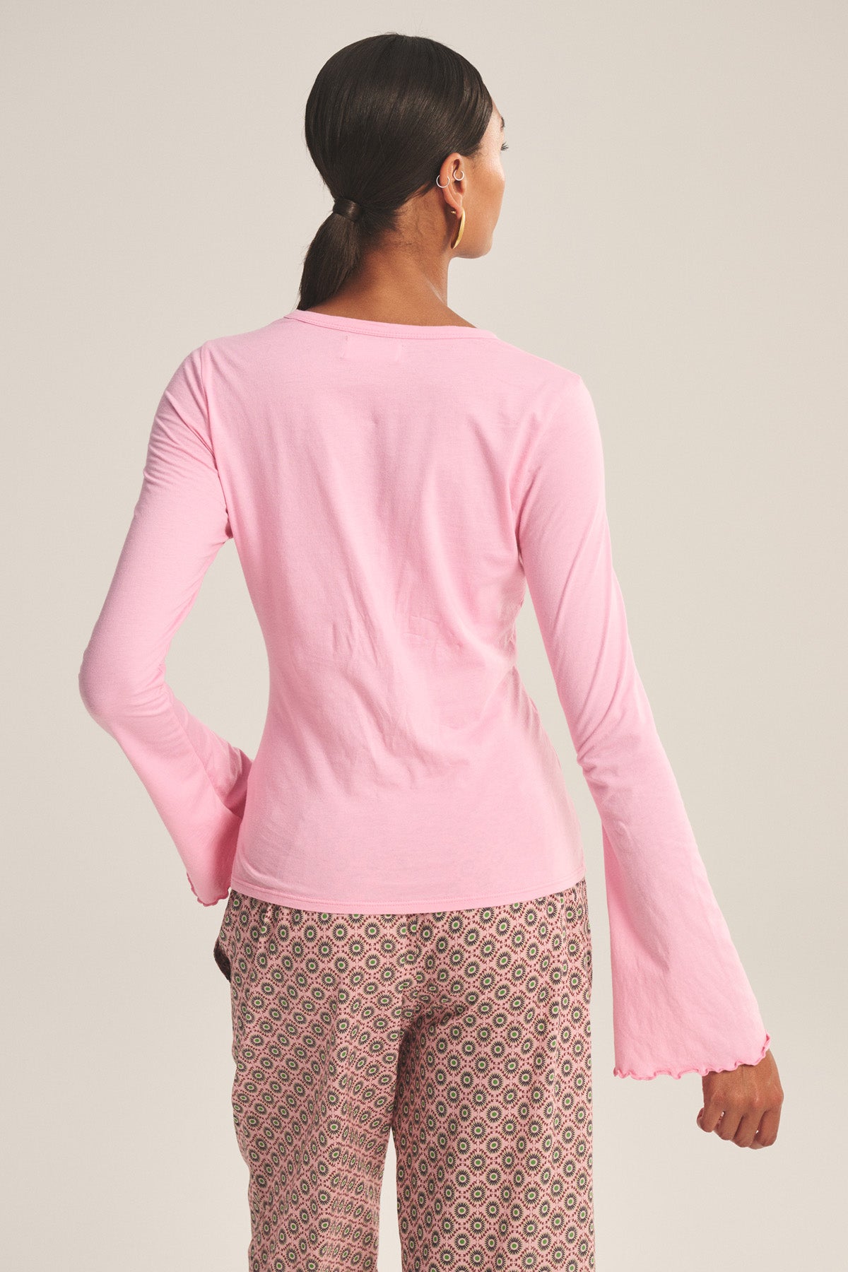 A person with dark hair in a low ponytail is seen from the back wearing the CAMILLE TEE by Velvet by Graham & Spencer—a long-sleeve pink top with scalloped cuffs—paired with patterned pants, set against a neutral background.