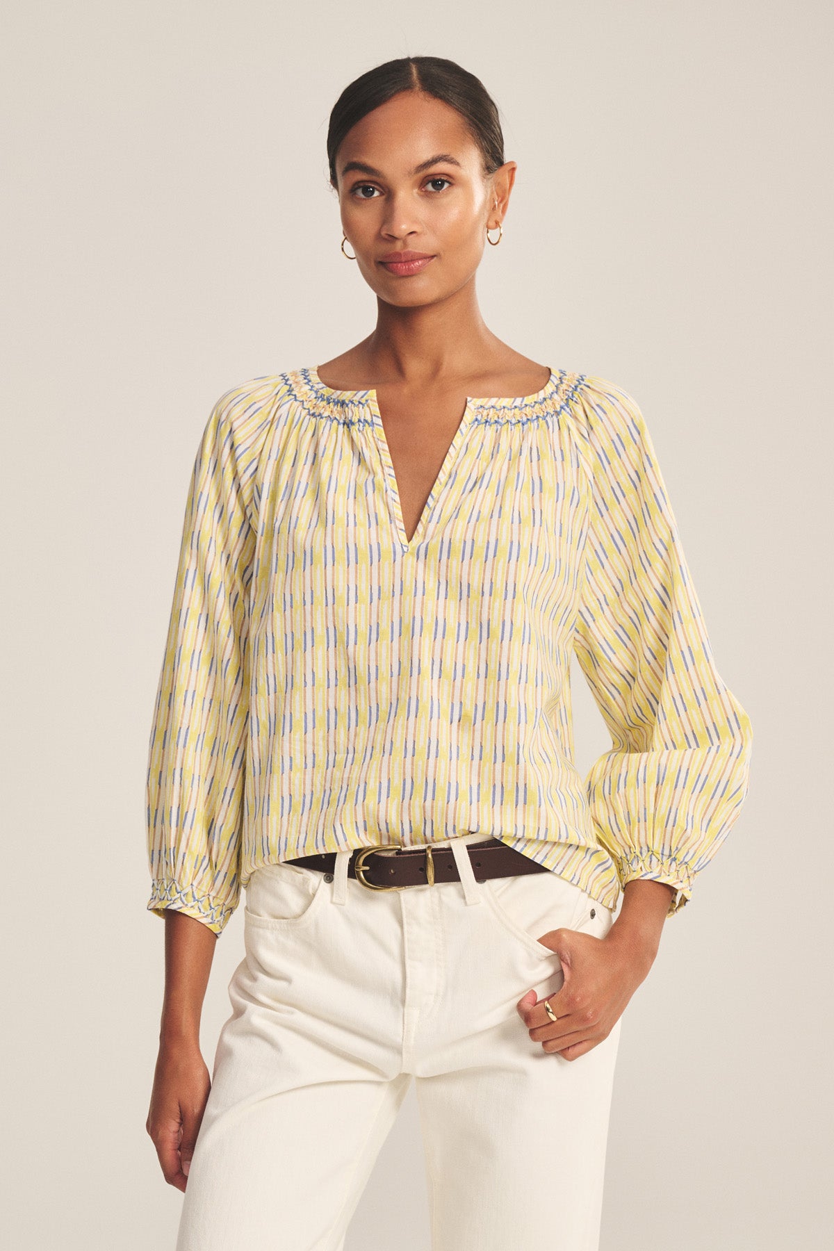 A woman wears the CAMPBELL TOP by Velvet by Graham & Spencer, a relaxed-fit cotton blouse with yellow and blue stripes and a V-neckline, styled with white pants against a neutral background.-41128063959233