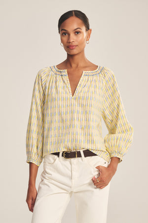 A woman wears the CAMPBELL TOP by Velvet by Graham & Spencer, a relaxed-fit cotton blouse with yellow and blue stripes and a V-neckline, styled with white pants against a neutral background.