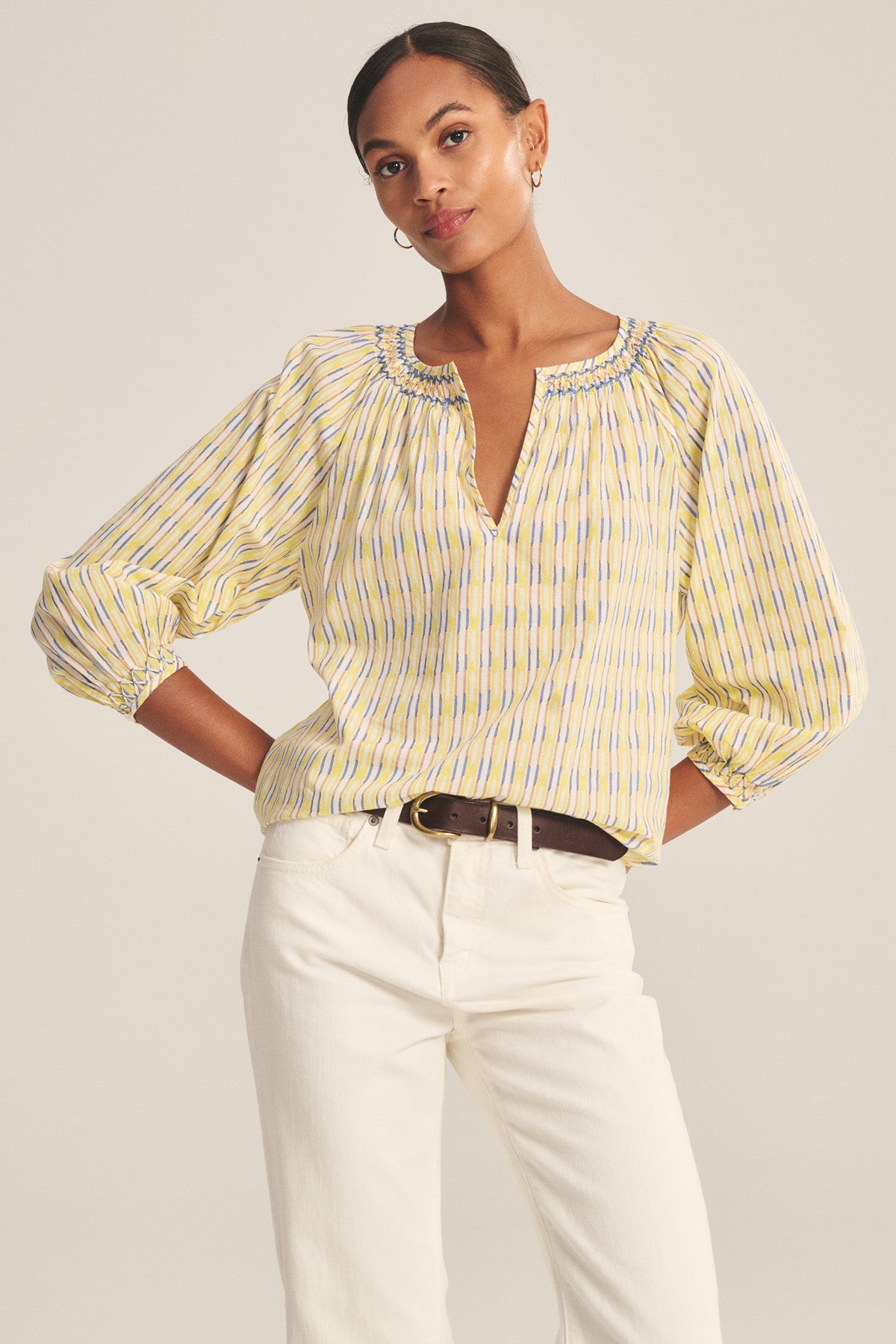 A woman wearing the Velvet by Graham & Spencer CAMPBELL TOP, a yellow and blue striped cotton piece with a gathered neckline, pairs it with white pants and stands with her hands on her hips.-41128063926465