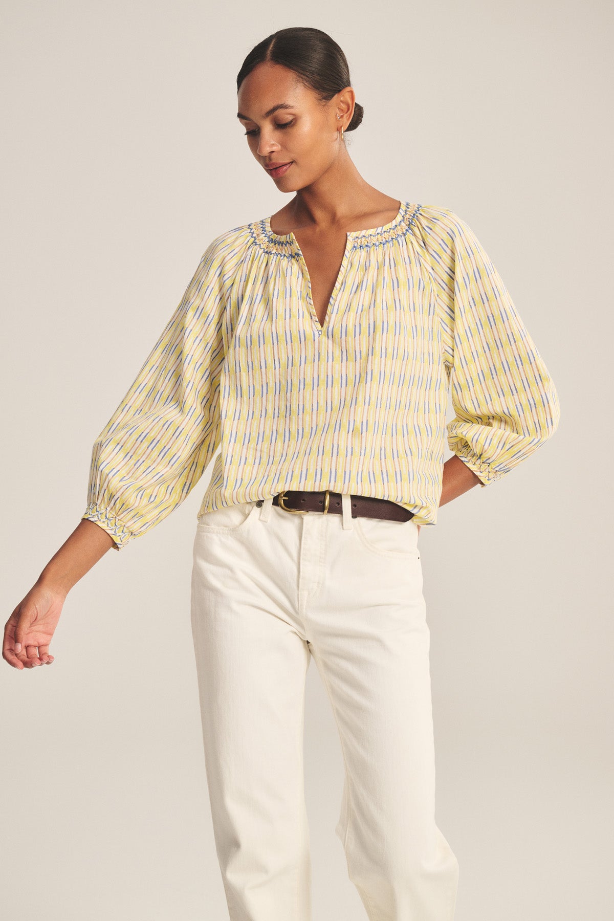 A person wears the CAMPBELL TOP by Velvet by Graham & Spencer, featuring yellow and blue stripes, balloon sleeves, a V-neckline, and white pants, standing against a plain background.-41128063893697