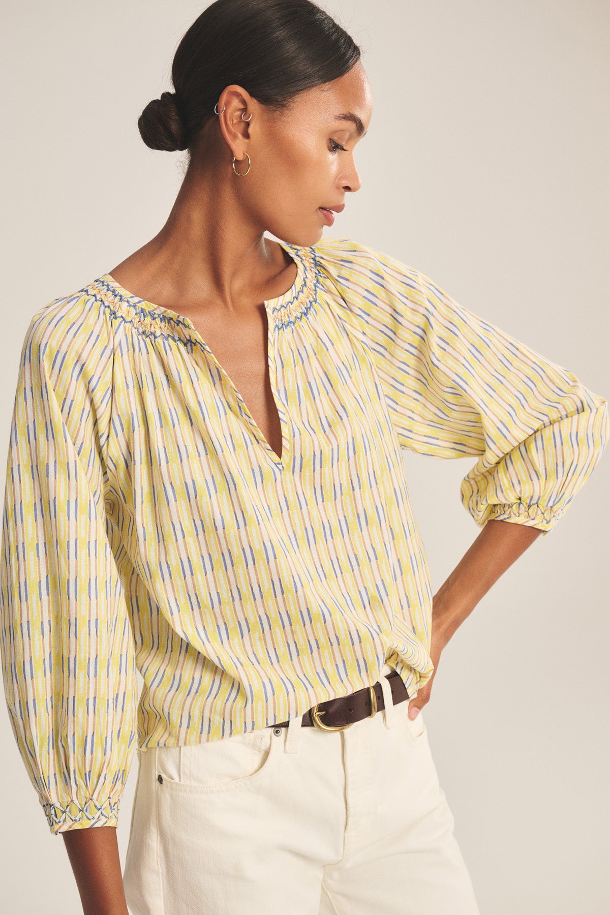 A woman stands confidently with one hand on her hip, wearing the CAMPBELL TOP by Velvet by Graham & Spencer—a relaxed cotton top featuring a smocked neckline and subtle yellow and blue stripes—paired with white pants and a brown belt.