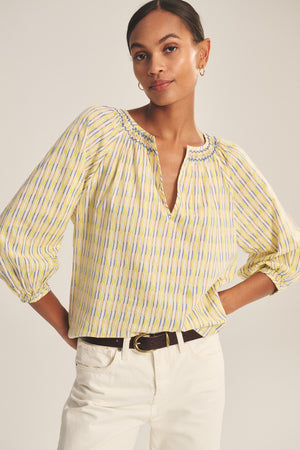 Woman wearing the CAMPBELL TOP by Velvet by Graham & Spencer—a yellow printed cotton top with a relaxed silhouette, V-neckline, and three-quarter sleeves—paired with white pants and a black belt, standing with hands on hips.