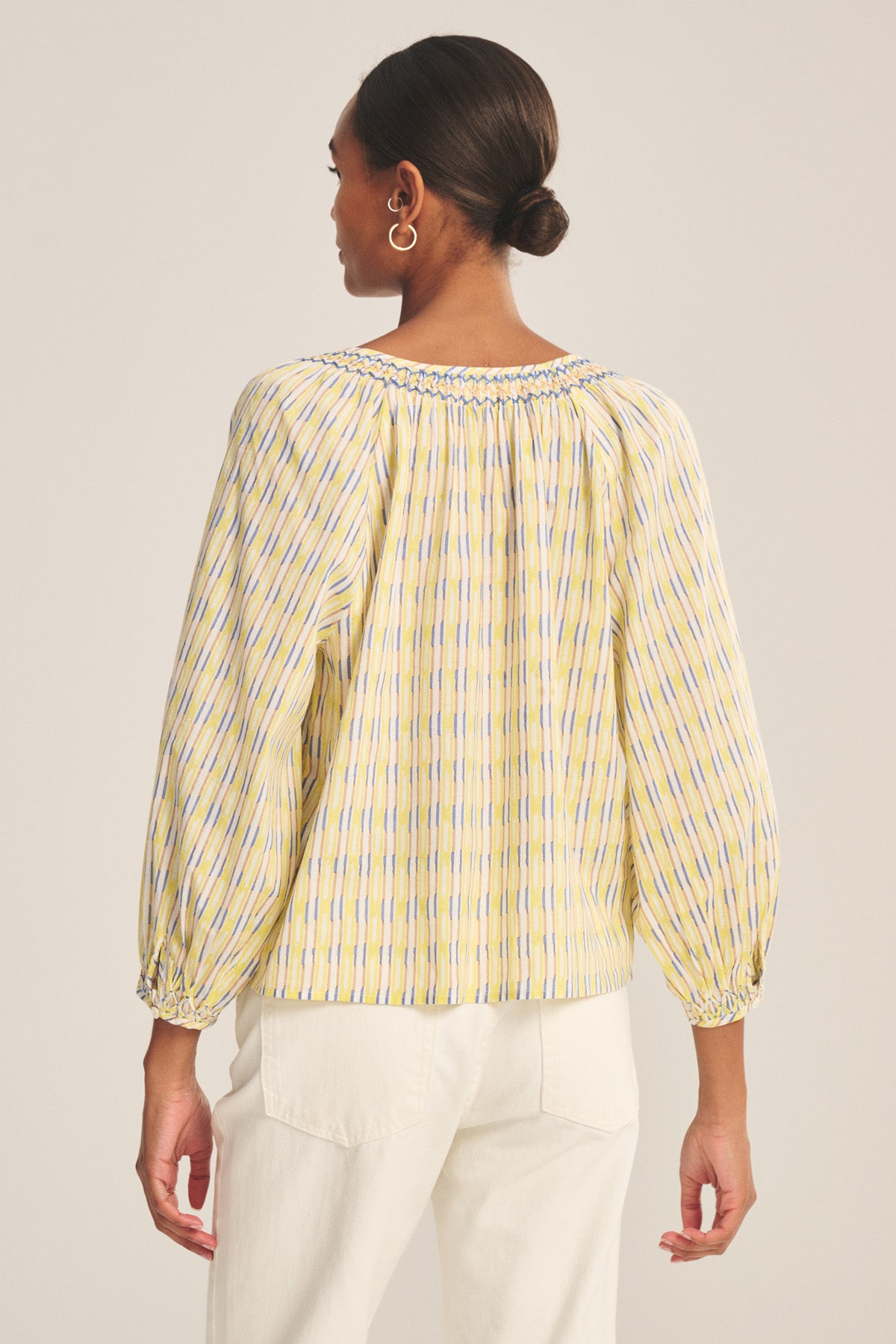 A woman with hair in a bun is shown from the back wearing the CAMPBELL TOP by Velvet by Graham & Spencer—a loose, long-sleeve, yellow and blue printed cotton top with a relaxed fit—paired with white pants.