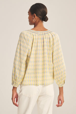 A woman with hair in a bun is shown from the back wearing the CAMPBELL TOP by Velvet by Graham & Spencer—a loose, long-sleeve, yellow and blue printed cotton top with a relaxed fit—paired with white pants.