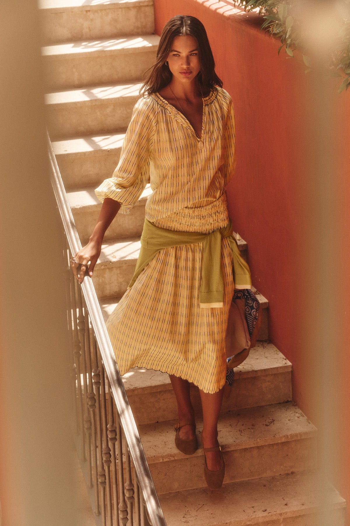 A woman wearing the CAMPBELL TOP by Velvet by Graham & Spencer and brown shoes stands on a staircase with a red wall, looking at the camera.-41128063664321