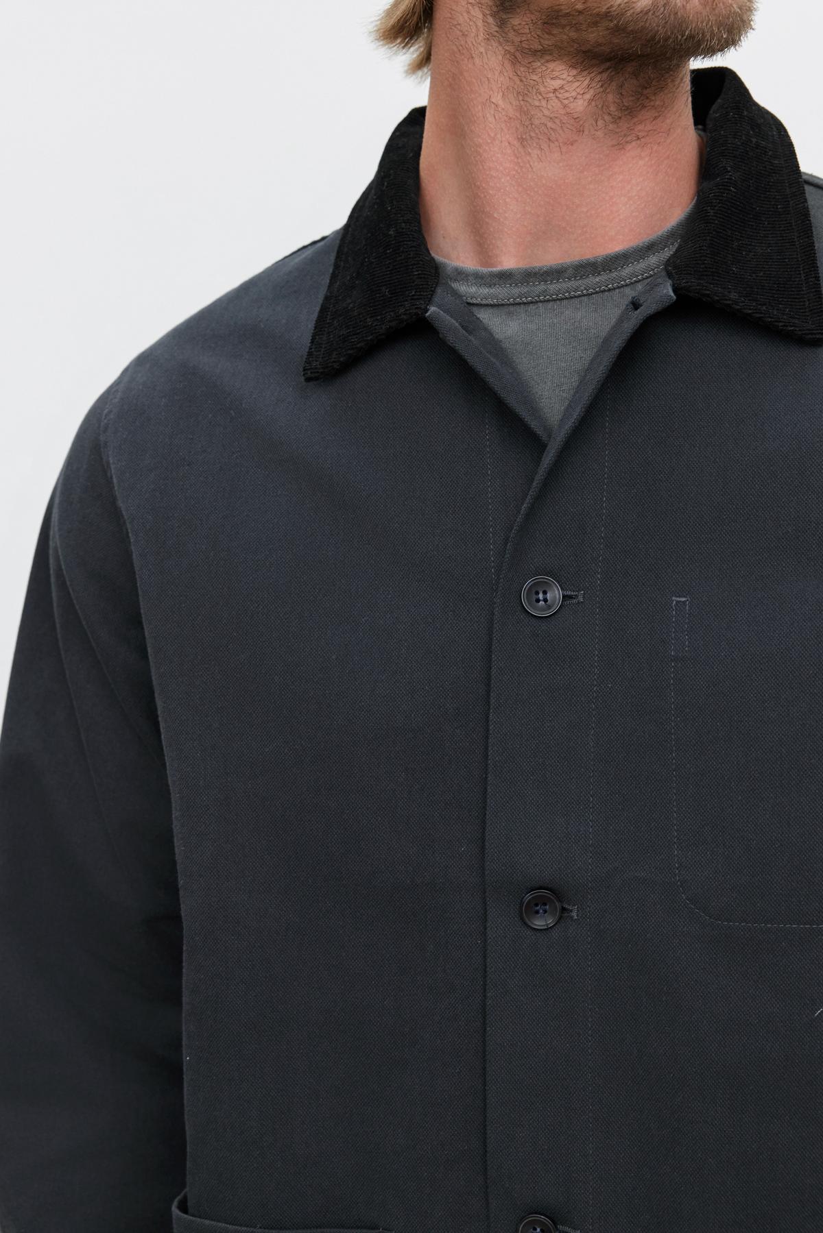   A person is wearing the EMORY JACKET from Velvet by Graham & Spencer, a structured dark-colored cotton canvas jacket with a corduroy collar, over a muted gray shirt. The image is cropped at the shoulders and chin, partially obscuring the face. 