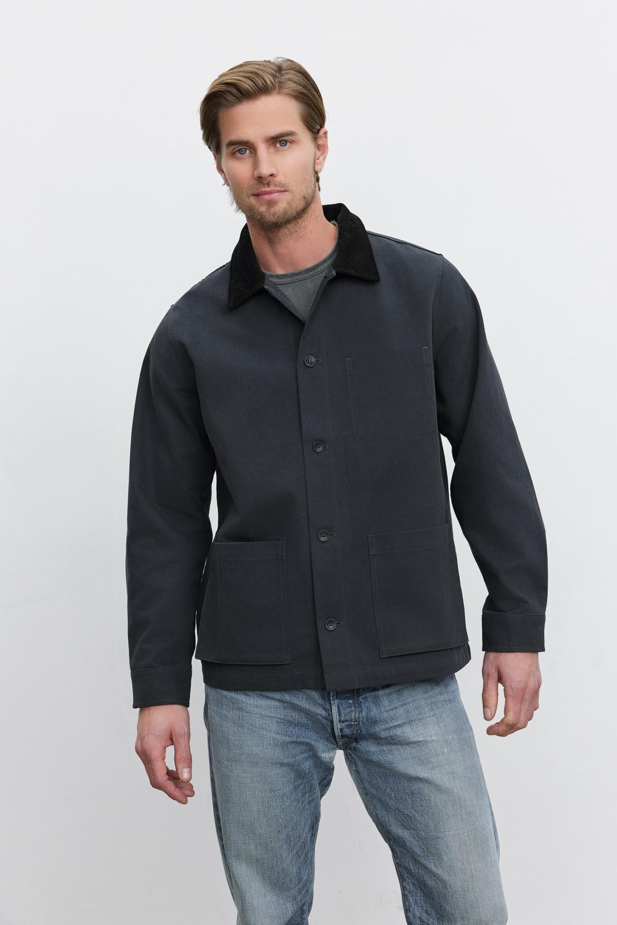   A man in an EMORY JACKET by Velvet by Graham & Spencer, featuring structured black cotton canvas with a corduroy collar, and light blue jeans stands against a plain white background. 