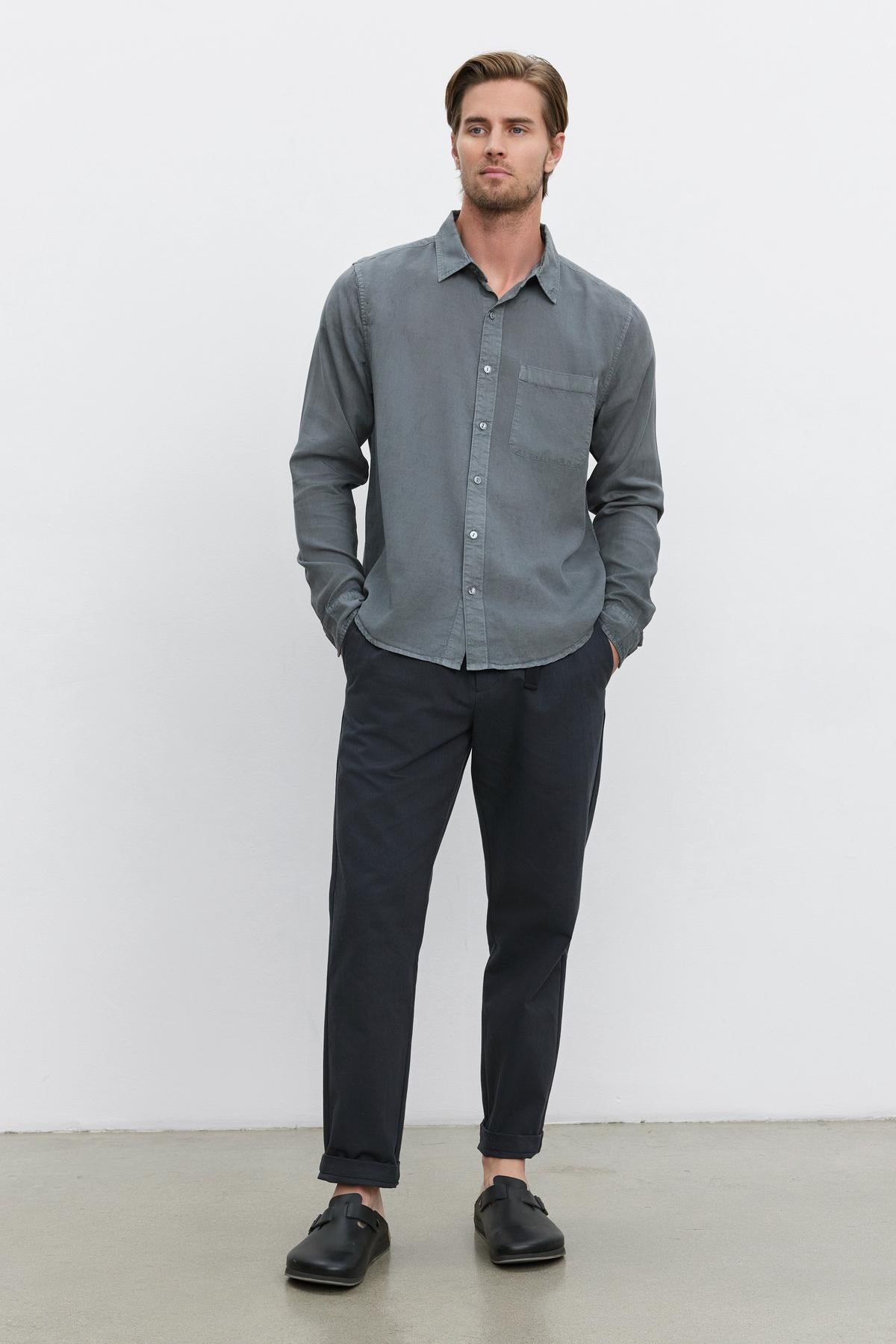   A man stands against a plain white background, hands in pockets. He is wearing a grey button-up shirt made from cotton canvas fabric, **MORAN PANT** in dark grey with an elastic waist by **Velvet by Graham & Spencer**, and black loafers. 