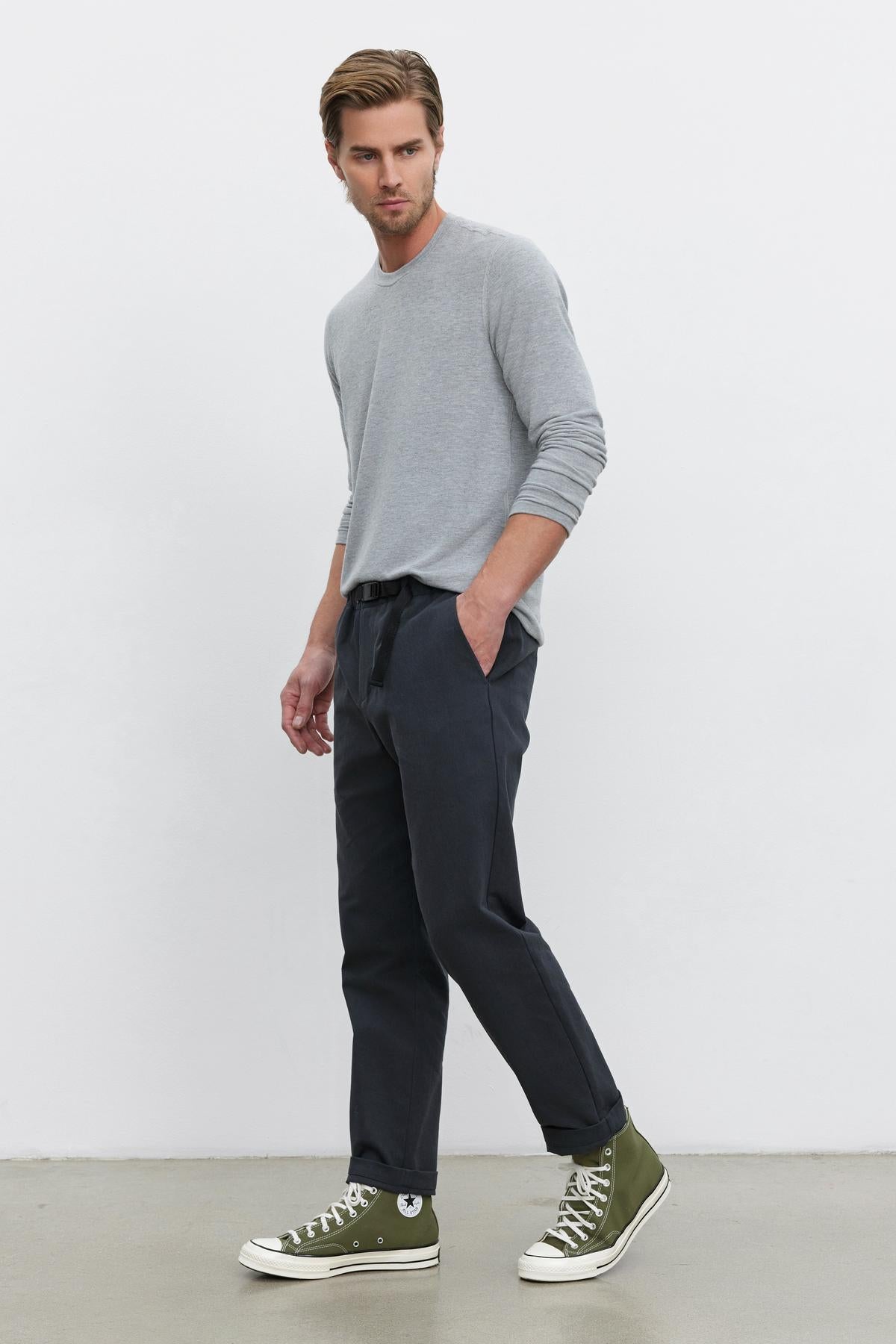 A man is standing against a plain white background, wearing a grey long-sleeve shirt, Velvet by Graham & Spencer's MORAN PANT with a tapered ankle design in dark color, and green high-top sneakers. His left hand is in his pocket, and he appears to be glancing to the side.-37648618815681