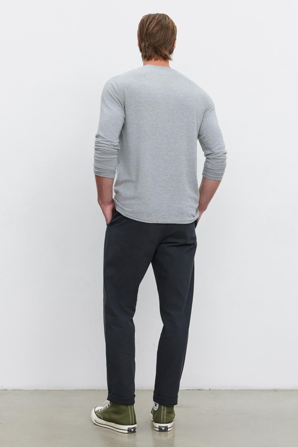   A person with light brown hair is standing with their back to the camera, wearing a grey long-sleeve shirt, the MORAN PANT by Velvet by Graham & Spencer paired with a woven belt, and green high-top sneakers, in front of a white background. 