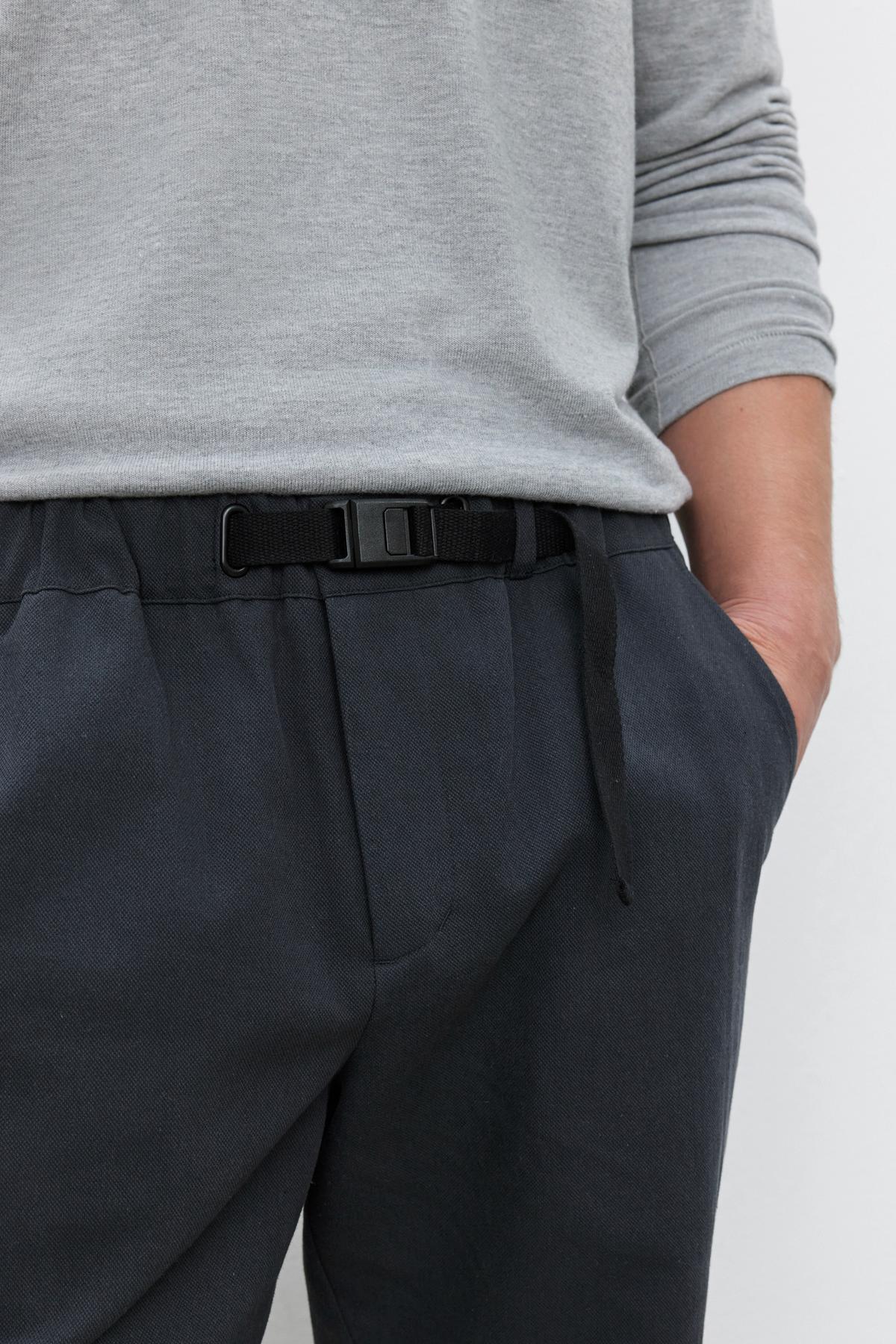   A person wearing a light gray long-sleeve shirt and Velvet by Graham & Spencer MORAN PANT in dark blue, with tapered ankles, their hands in their pockets, featuring a black woven belt with a buckle. 