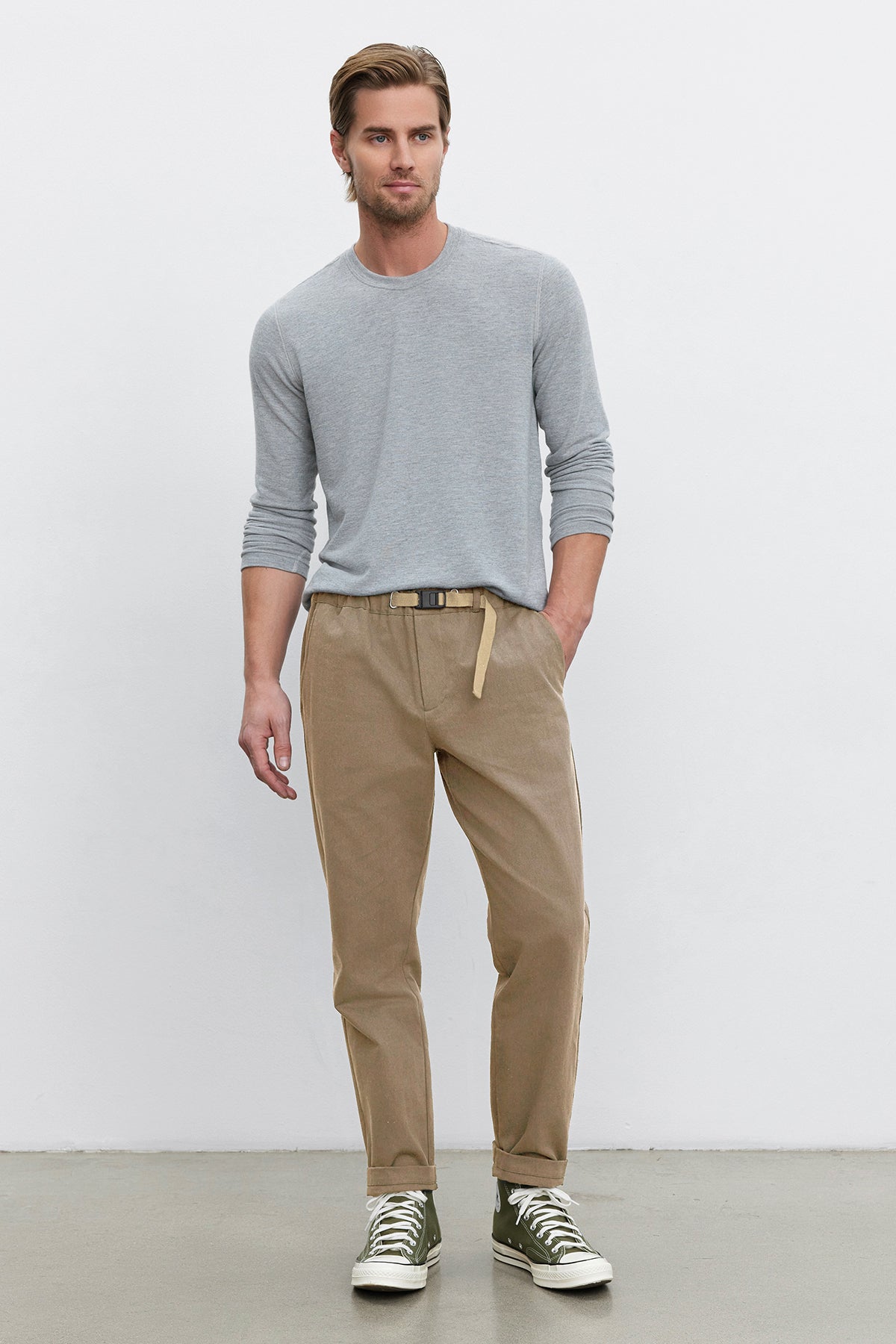   A man wearing a light gray long-sleeve shirt crafted from cotton canvas fabric, MORAN PANT by Velvet by Graham & Spencer with a matching belt, and green sneakers stands against a plain white background. 