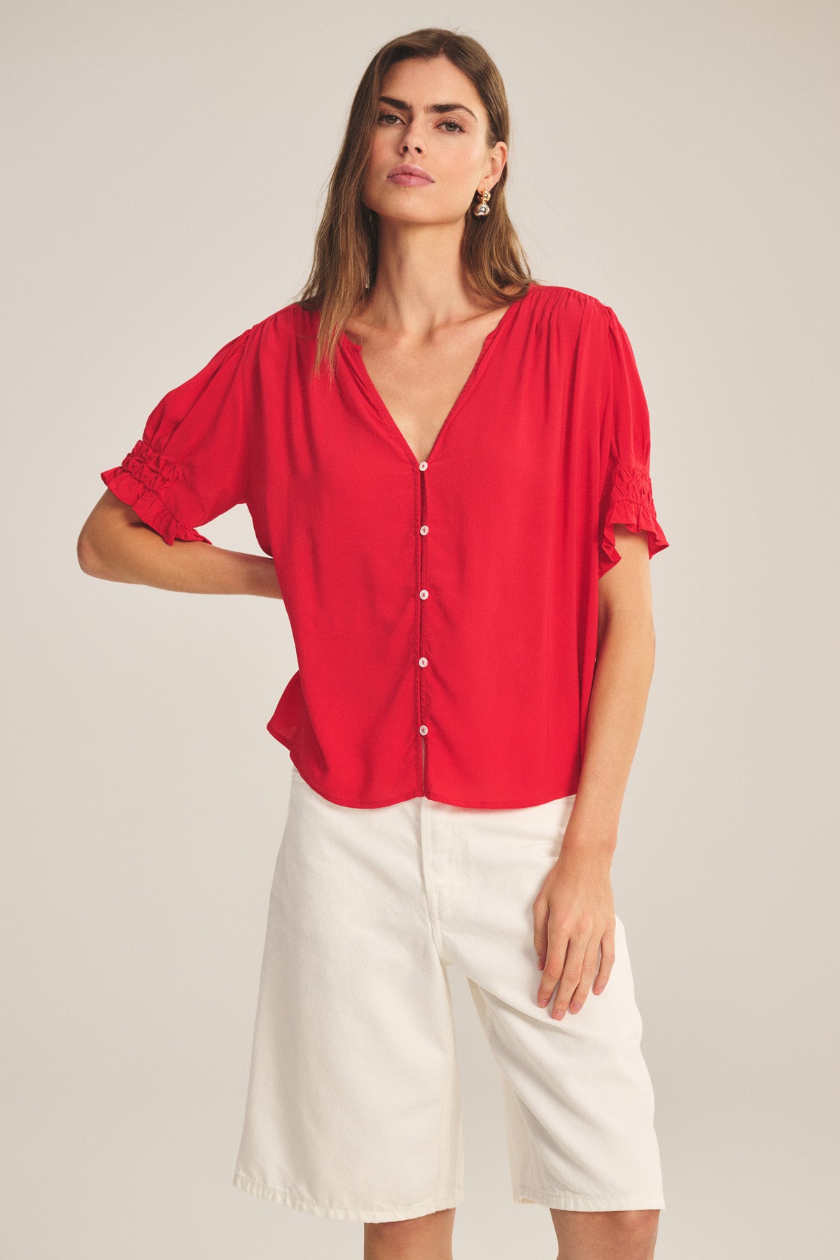 A woman models the Velvet by Graham & Spencer CARMELLA TOP, a red V-neck button-front blouse, paired with white knee-length shorts, standing against a plain background and facing the camera.-41081213026497