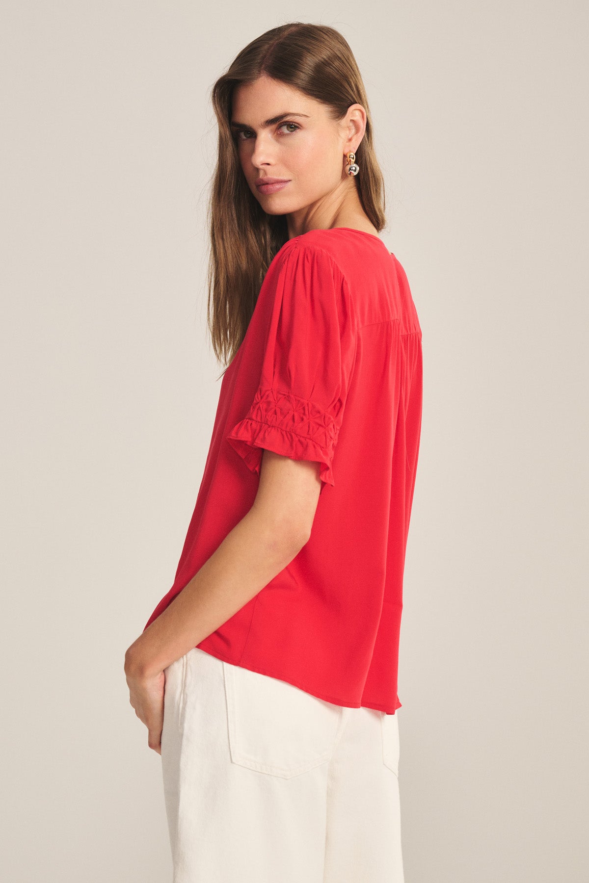 A woman stands sideways, looking over her shoulder, wearing the CARMELLA TOP by Velvet by Graham & Spencer—a red V-neck, button-front blouse with embroidered short sleeves—paired with ivory pants.