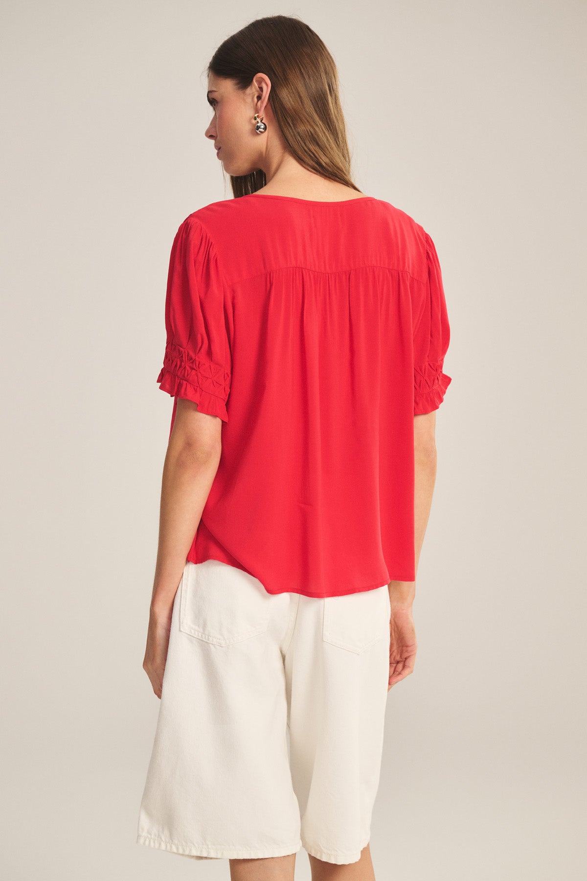 A woman faces away, wearing the CARMELLA TOP by Velvet by Graham & Spencer—a red flowy blouse with gathered details—paired with loose white knee-length shorts.-41081212928193