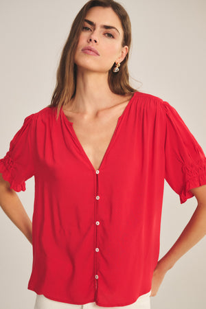 A woman models the CARMELLA TOP by Velvet by Graham & Spencer, a loose red rayon challis blouse with short puff sleeves, white buttons, and a deep V-neck, standing against a plain background.