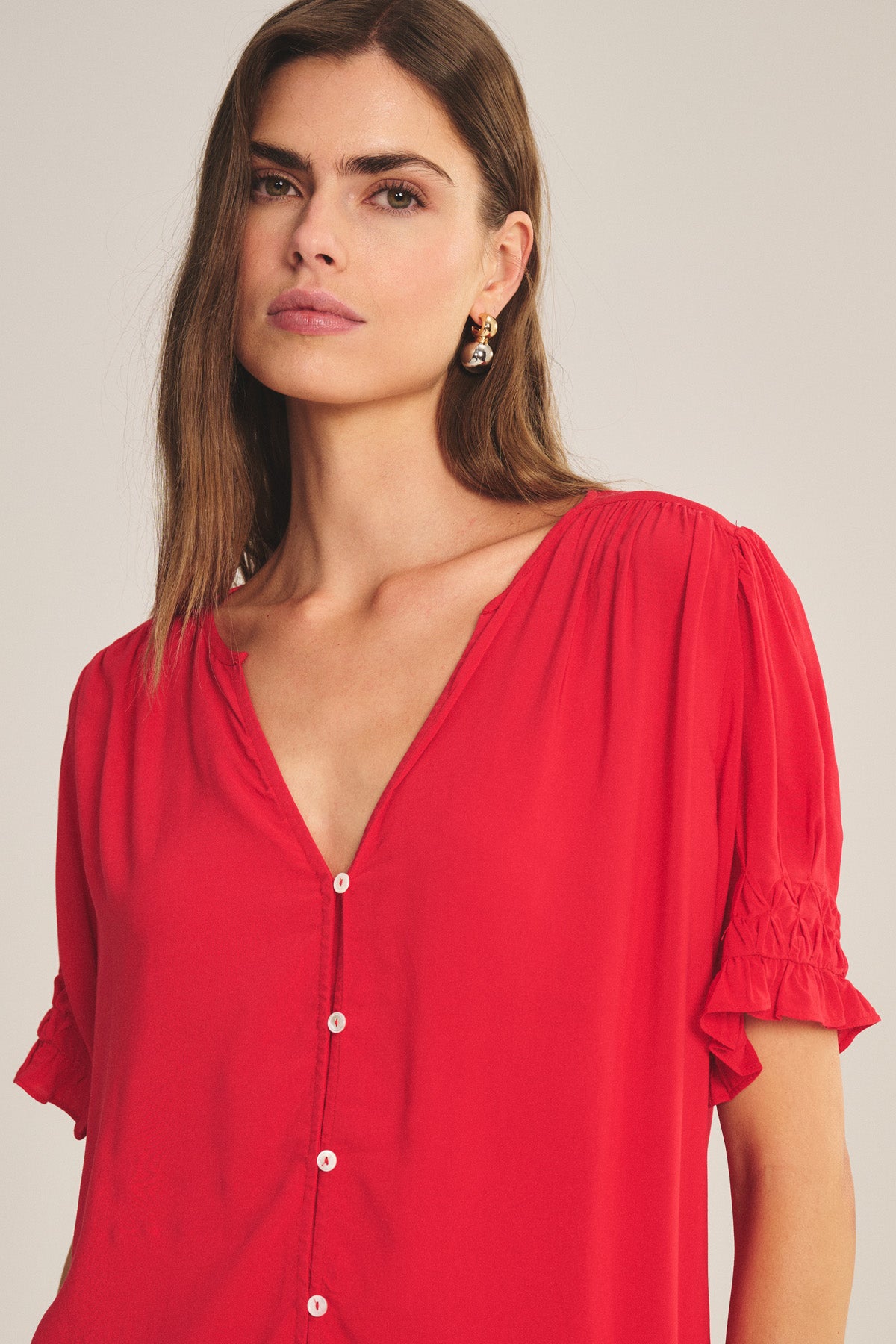 Woman with long brown hair wears the Velvet by Graham & Spencer CARMELLA TOP—a red V-neck, button-front blouse with short ruffled sleeves and a flowy fit—looking directly at the camera against a plain background.-41081212862657