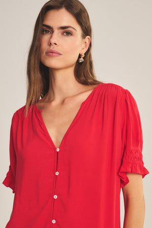 Woman with long brown hair wears the Velvet by Graham & Spencer CARMELLA TOP—a red V-neck, button-front blouse with short ruffled sleeves and a flowy fit—looking directly at the camera against a plain background.