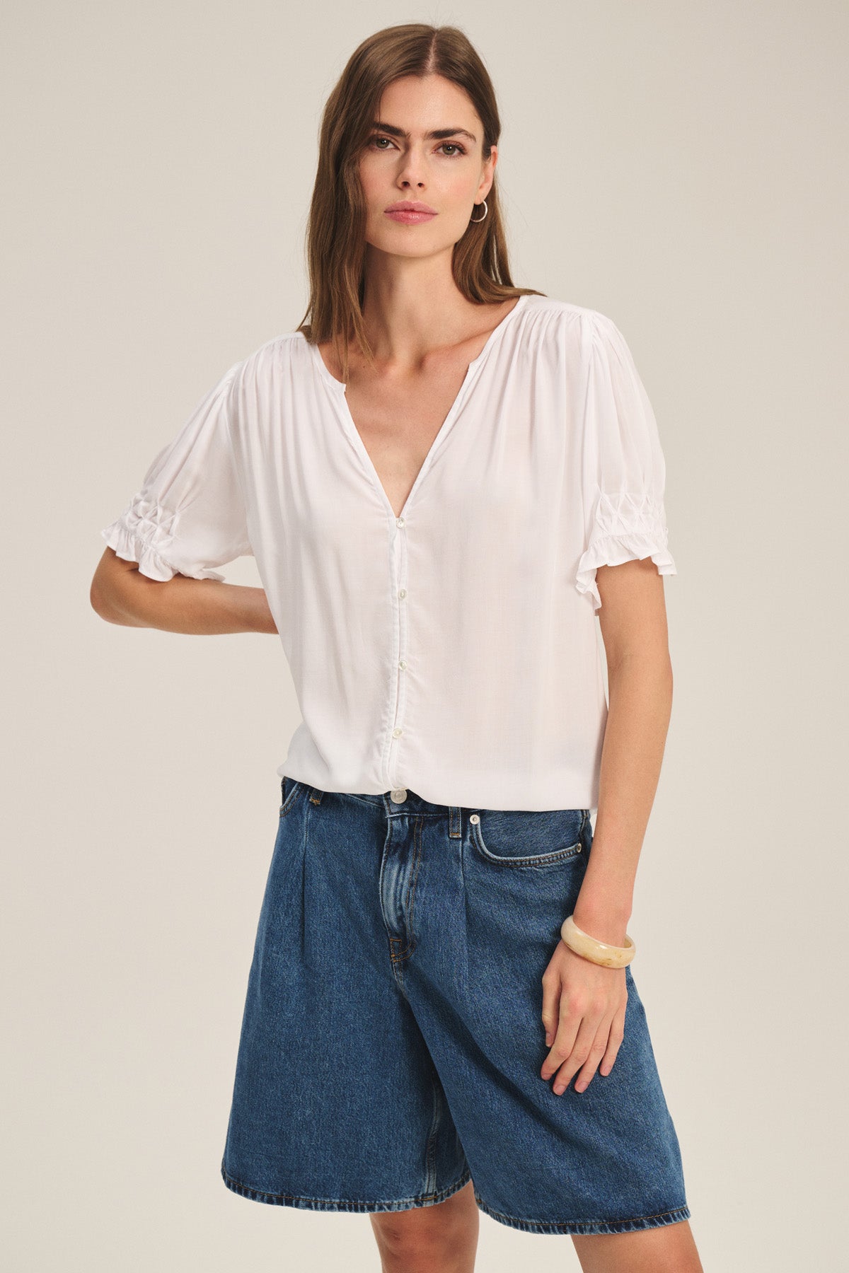 A woman stands against a plain background wearing the CARMELLA TOP, a white V-neck button-front blouse by Velvet by Graham & Spencer, paired with loose blue denim shorts.