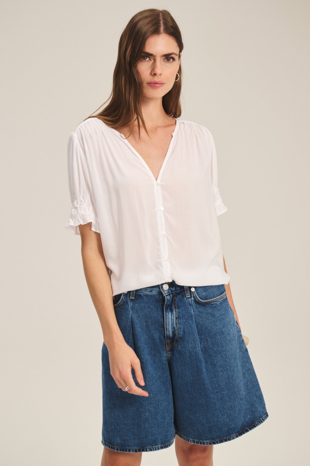 A woman models the CARMELLA TOP by Velvet by Graham & Spencer—a flowy V-neck blouse—paired with high-waisted blue denim shorts, standing against a plain light background.